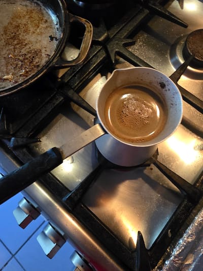 A stovetop in an indoor kitchen in Tuapse, Russia, shows a partially visible old cast iron pan with remnants of food in it, and a small metal pot filled with dark liquid, likely coffee. The pot has a black handle and is placed on the gas burner. The stovetop appears to be made of stainless steel and is showing signs of use and some light residue. The lighting suggests it might be daytime, with artificial light also contributing to the reflections on the metal surfaces. The overall impression is a utilitarian kitchen environment.