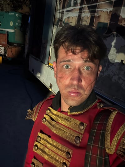 A close-up, medium shot captures a person wearing a red military-style jacket with gold trim and buttons, and a black collar. The individual's face is visible, showing striking blue eyes, a slightly open mouth, and visible makeup or prosthetics resembling scars on their cheeks and forehead. The person is looking directly at the camera. The background is dimly lit and appears to be outdoors, with a large, weathered vehicle or structure behind them. Visible objects in the background include a green generator and what looks like a piece of discarded material with tear marks. The overall lighting suggests it might be nighttime or a dark, indoor set. There is no discernible information about the location being Gollagutta, India.