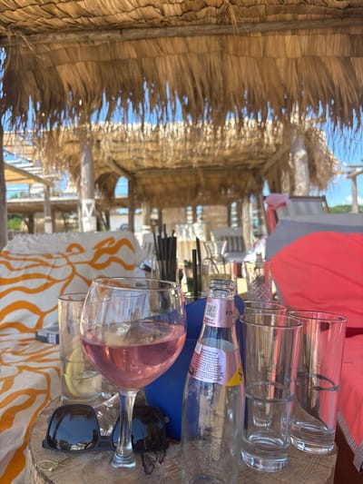 The image depicts a close-up view of a wooden table at a beachside establishment in Shkorpilovtsi, Bulgaria. Overhead, a thatched roof provides shade, with glimpses of a blue sky and distant buildings visible through its gaps. On the table, there are several drinking glasses, a wine glass filled with rosé wine, a partially empty bottle of Schweppes Pink Pepper Tonic, and a pair of black sunglasses. To the left, a white cloth with orange wavy stripes is partially visible. In the background, other tables, seating, and beach umbrellas suggest a social gathering area. The scene appears to be set during daylight hours.