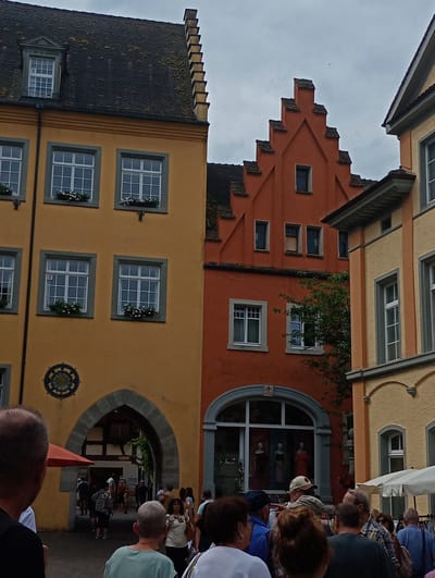 In Meersburg, Germany, this image captures a view of a street with historic buildings and pedestrians. The foreground shows the backs of several people, with one person wearing a black t-shirt and another in a light blue shirt. In the middle ground, an arched gateway is visible on a yellow building, with people walking through it into a shaded area. The gateway is adorned with a circular decorative element above it. Adjacent to the yellow building is an orange building with a distinctive stepped gable. Further back, more people are seen browsing at stalls beneath a striped awning. The sky is overcast. The buildings exhibit traditional European architecture with multiple windows, many featuring flower boxes.