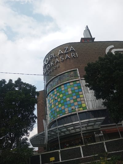 A large commercial building, likely a shopping mall or plaza, dominates the center of the frame, viewed from a slightly low angle looking upwards. The building has a distinct curved facade, giving it a cylindrical or rounded appearance. The upper section of the building is clad in a patterned, brownish-tan material, possibly tiles or panels, with a large white sign spelling out "PLAZA LOKASARI" in prominent capital letters. Below "LOKASARI," the word "BOGOR" is written in a smaller font, indicating the location.
Above the main sign, on the building's roof, there's a distinctive, pointed architectural feature, appearing metallic or grey. Below the main sign, a significant portion of the building features a large, curved section with numerous square panels arranged in a checkered pattern of varying shades of blue, light blue, green, and yellow. This section likely represents large windows or a decorative wall.
Further down, towards the base of the building, there are large glass windows, suggesting storefronts or dining areas. A white, modern-looking awning or overhang extends over these lower sections. On the ground level, a dark, textured wall, possibly stone or concrete, forms the base. A sign for "Solaria" is visible on one of the lower storefronts.
Large, lush green trees frame the building on both the left and right sides, with their dark foliage partially obscuring parts of the structure, particularly the left side and some of the right. The sky is overcast with a mix of white and light grey clouds, indicating a cloudy or partly cloudy day. A black power line or cable stretches horizontally across the upper left quadrant of the image. The overall setting appears to be an urban or suburban commercial area during daylight hours.