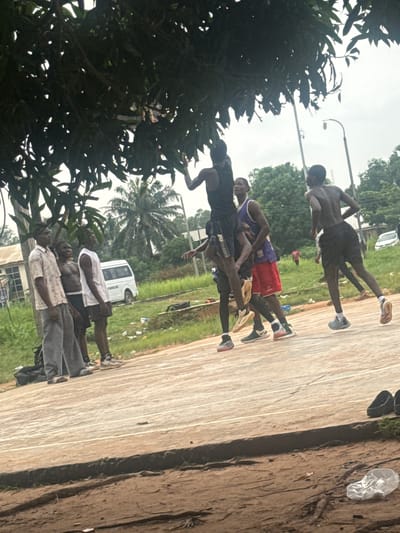 In Amansee, Nigeria, on a cloudy day, a group of young men are engaged in a basketball game on an outdoor court. The court surface appears to be worn and cracked concrete with painted lines. Lush green trees with dense foliage overhang parts of the scene, casting shadows. In the background, there are buildings, a white van, and a lamppost, suggesting an urban or semi-urban setting. The players are dressed in casual athletic wear, with some barefoot and others in sneakers. One player in a black tank top is captured mid-air, reaching for a basketball, while others are positioned around him, observing or actively participating in the game. The overall atmosphere seems to be one of casual recreation and community.