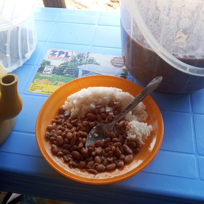 The image shows a plate of rice and beans on a blue plastic table in Borabu Chitago ward, Kenya. A silver spoon rests on the plate. Beside the plate are a clear plastic container filled with liquid and a yellow dispenser. A laminated advertisement for "ZPL Coffee / Sits" is visible on the table.