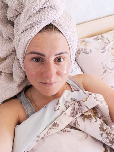 A woman with blonde hair wrapped in a towel and wearing eye patches lies in bed. She is wearing a grey tank top and a floral print comforter. The background shows a pillow with a floral pattern. The image is a close-up, top-down view of the woman.
