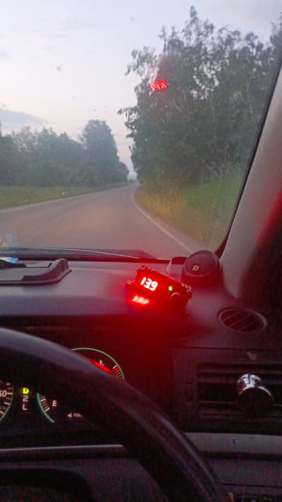 The image shows the interior of a car driving on a road with trees on either side. The dashboard has a red digital display showing "13.9". There are also instrument cluster gauges visible, including a tachometer with a red needle pointing upwards and a fuel gauge. The sky is overcast. The location is identified as Zuy, Russia.