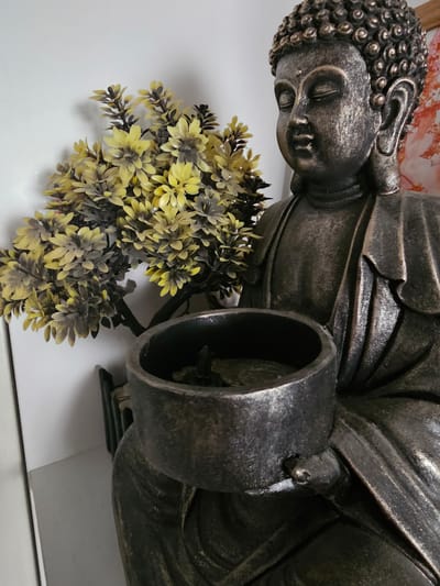 A close-up, low-angle, sideways shot features a statue of a sleeping Buddha, rendered in a dark, metallic finish with antique bronze accents. The statue is positioned horizontally, with the Buddha's head on the left and the body extending to the right. The Buddha's eyes are closed, conveying a sense of peace and tranquility.
In the foreground and partially beneath the statue, a cluster of artificial flowers with yellow and dark grey petals adds a touch of artificial greenery. The flowers appear to be made of plastic or silk, with detailed petals and dark stems.
The background is largely out of focus, but hints of a textured, possibly floral or abstract pattern in shades of pink and orange are visible behind the Buddha's head, suggesting a decorative element or artwork. The setting appears to be indoors, with a smooth, white surface beneath the statue and flowers, reflecting light and creating a soft shadow.
The lighting is even and diffused, suggesting an indoor environment with artificial light. No specific time of day or weather conditions can be inferred. No text is visible in the image. The overall impression is one of serene decoration, possibly within a home or a place of worship.