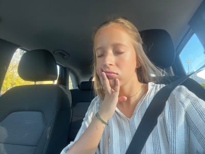 A young female occupies the front passenger seat of a vehicle, in Aldoar, Foz do Douro e Nevogilde, Portugal, during daylight hours. She has light-colored hair, closed eyes, and her right hand supports her chin/jaw. She wears a light-toned, vertically striped top, a thin dark necklace, and a green bracelet on her left wrist. A black seatbelt diagonally crosses her torso. The car's interior is dark-toned, with a black headrest visible to her left. Through the driver's side window, bright green-yellow foliage is visible. The passenger's side window reveals a clear blue sky, a distant street light pole, and a partial building structure.