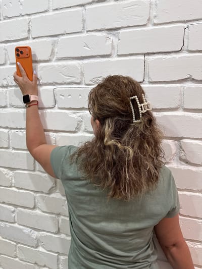 The media file shows a woman holding an orange smartphone against a white brick wall. The woman is wearing a light green t-shirt and has curly brown hair adorned with a pearl hair clip. On her left wrist, she wears a pink smartwatch. The background is a textured white brick wall with visible cracks and grout lines. The overall scene appears to be indoors, with neutral lighting, suggesting it could be daytime. The woman's pose and the way she holds the phone suggest she is taking a picture or video. There are no discernible emotions or specific weather conditions indicated. The location context provided is "Столбище, Russia". No visible text is present in the image.