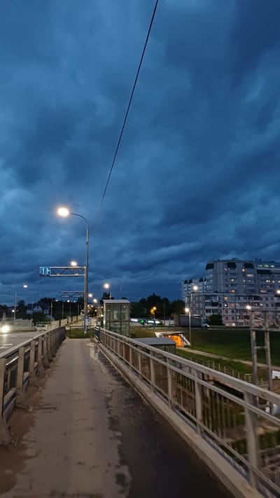 The image depicts a nighttime scene on a bridge in Obukhovo, Russia. The view is oriented sideways, with the city skyline and a dramatic, cloudy sky on the left side of the frame. A large apartment building with illuminated windows is visible in the distance. Streetlights line the bridge, casting a warm glow on the wet pavement and metal railings. A power line stretches across the sky. A bus stop sign is also visible. There are no people visible in the image. The focus is on the urban landscape at dusk.