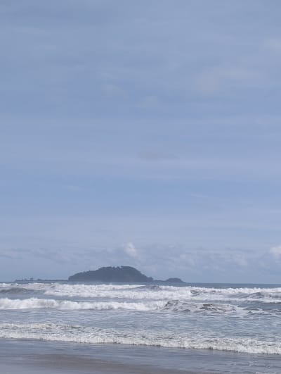 This is a photograph taken at Meudheun Beach, Aceh Province, Indonesia. The image captures a serene coastal scene with several layers of ocean waves rolling towards the shore. In the distance, a small, forested island is visible on the horizon, adding a sense of depth to the landscape. The sky above is a pale blue, with some scattered clouds, suggesting fair weather. The foreground shows the wet sand of the beach. There are no people or man-made structures visible in the frame. The overall impression is one of natural beauty and tranquility.