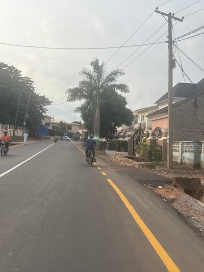 This is a vertically oriented, low-angle shot of a street scene in Kugbo, Nigeria. The road, freshly paved with a distinct yellow line down the center, curves gently to the right. On the left side of the road, a tall utility pole with numerous wires stretches upwards against a pale, overcast sky. Beside the pole, a large palm tree with feathery fronds dominates the mid-ground, its fronds silhouetted against the bright sky. Behind the palm tree, several multi-story buildings with light-colored facades and visible windows are lined up. Some of these buildings feature decorative ironwork in front, hinting at an ornamental style. To the right of the road, a raised embankment of reddish-brown earth and rocks marks the edge of the paved area.
Several people on motorcycles are visible on the road. In the foreground, two individuals are riding motorcycles side-by-side. Further down the road, a person on a motorcycle is moving away from the camera, and another motorcycle can be seen further still. A white car is parked near the buildings on the left. The overall impression is a bright, possibly early morning or late afternoon scene, with diffused light suggesting cloudy weather. The presence of utility poles and the style of the buildings suggest a moderately developed urban or suburban area. No visible text is present in the image.