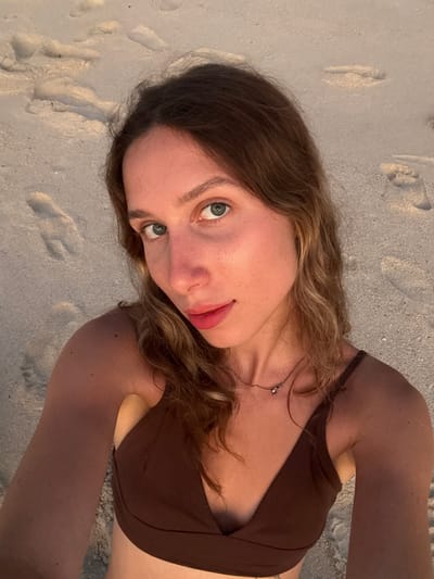 The image shows a close-up, high-angle selfie of a young woman with blonde hair and blue eyes, taken on a sandy beach. She is wearing a brown bikini top and a delicate necklace. Her face is turned slightly to her left, and she is looking directly at the camera. The background is composed of sand with visible footprints. The lighting suggests it is either sunrise or sunset, casting a warm glow on the scene. The woman's skin has a tan appearance, and there are a few freckles visible on her nose and cheeks. The overall setting is a beach in Kertasari, Indonesia.