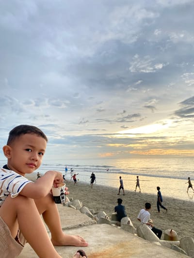 A young boy with short hair is sitting on concrete barriers on a beach in Bateetimoh, Indonesia, holding a cup of ice cream. He is wearing a striped t-shirt and tan shorts. In the background, several people are playing soccer on the sandy beach near the ocean. The sky is filled with clouds, with the sun setting over the water, casting a warm glow. The scene depicts a relaxed evening at the beach.
