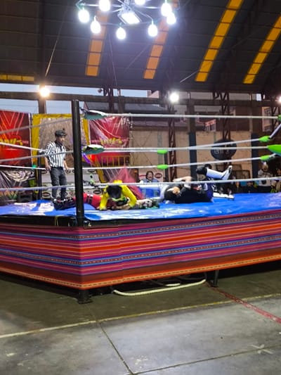 A wrestling match unfolds inside a ring adorned with colorful striped fabric. Two wrestlers are down on the blue mat. A referee in a striped shirt stands near the corner, observing. The ring is surrounded by ropes and turnbuckles. Background shows spectators, banners, and the building's structure with visible support beams and a metal roof, illuminated by overhead lights.