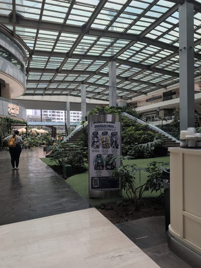 The image depicts an interior view, likely of a shopping mall or commercial space, in Jesús del Monte, Mexico. The architecture features a large glass ceiling supported by metal framework, which allows for ample natural light. An elevated walkway or second level is adorned with greenery, including various plants and artificial turf. A sign in Spanish, advertising an after-school program with the words "UN ESPACIO AFTER SCHOOL DISEÑADO PARA" and further text detailing "CREATE", "BUILD" and "EXPRESS", is prominently displayed. The floor is divided into dark and light sections. A person is walking on the darker section of the floor. Store signs are visible in the background, including "AMERICAN OUTLET" and "DEFENSA PERSONAL."