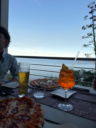 A close-up, eye-level shot shows a table set for a meal with a picturesque view of the sea and sky. In the foreground, a pepperoni pizza is partially visible, while a second pizza with mushrooms and cheese sits further back on the table. Two drinks are present: a tall glass of beer with foam and a vibrant orange cocktail garnished with a slice of orange and a white straw. A person, partially visible on the left, wears a striped shirt and sunglasses on their head. The table is covered with a dark-patterned tablecloth. In the background, a railing separates the table from the sea, which appears calm under a clear, pale blue sky. A tall, dark, spiky plant is visible on the right side of the frame, with the scene suggesting a coastal restaurant setting in Santa Maria Navarrese, Italy.