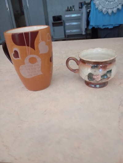 Two ceramic mugs sit on a light-colored, marbled surface. The mug on the left is a tall, orange mug with a dark brown handle. It is decorated with dark brown and white hearts, and the text "WITH LOVE FOR YOU" repeated in white. The mug on the right is smaller and wider, with a decorative brown glaze that fades to white. It features a painted pink flower with green leaves. The background is blurred, but appears to be an indoor setting with furniture and clothing visible, suggesting a domestic environment in Strășeni, Moldova.