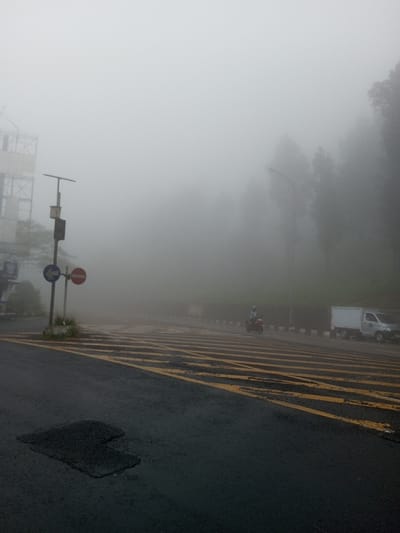 A foggy road scene with yellow stripes across the asphalt. A street sign with a no-entry symbol stands to the left. In the distance, a motorcyclist rides alongside a small white delivery truck. Trees and a building are visible through the thick fog. The overall atmosphere is muted and visibility is greatly reduced due to the heavy fog.