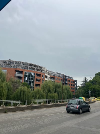 The image, taken in Sandanski, Bulgaria, shows a grey, overcast sky and a street scene with modern architecture and vehicles. In the foreground, a grey hatchback car is parked on the right side of the road, its license plate reading "E 7436 MK". Further down the road, two yellow taxis are visible.
The background features a contemporary building with a curved facade, clad in orange brick and large glass windows. The building appears to be under construction or recently completed, with exposed concrete elements visible in its structure. Lush green trees, possibly willows due to their drooping branches, surround the building and line the street, creating a natural contrast with the urban development. A metal railing separates the sidewalk from the greenery.
The overall impression is a quiet, somewhat muted urban environment under a cloudy sky. There are no people visible in the image. The time of day is not explicitly indicated but the overcast conditions suggest it could be daytime, perhaps morning or afternoon.