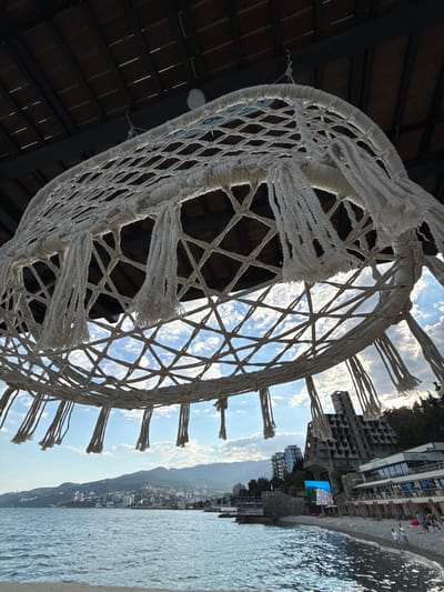 A low-angle, close-up shot focuses on a macrame hammock hanging from a dark, wooden roof. The hammock, made of thick, off-white rope, features a diamond-patterned net with tassels hanging from its lower edge. Through the hammock's open weave, a bright blue sky with scattered white clouds is visible, as well as a distant cityscape nestled against rolling green hills. The sea, a shimmering blue, laps at a pebble beach in the foreground. To the right of the frame, an unusual, brutalist-style concrete building with exposed structural elements looms over a modern-looking resort with a "Ski East" sign. Several beach chairs are set up on the shore, and a few people are walking or standing on the beach. The overall scene suggests a seaside resort in Yalta, Ukraine, on a bright, possibly late afternoon day.