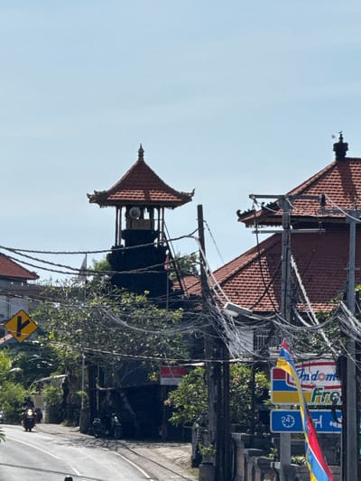 The image depicts a street scene in Kutuh, Indonesia, taken during the daytime. The sky is clear and light blue. A traditional Balinese structure with a tiered roof, possibly a temple or community hall, stands on the left side of the frame. Power lines are strung haphazardly between utility poles. On the right side of the image, commercial signs for "Indomaret" and "First" are visible, indicating the presence of a convenience store or a similar business. A triangular yellow street sign with a black arrow pointing left is present on the lower right. A road is visible on the right, with a motor bike driving away. Green foliage and trees fill the spaces in between buildings. The overall impression is of a typical busy street in a small town in Indonesia.
