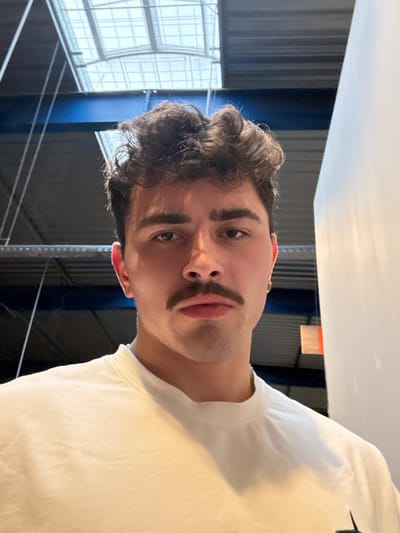 This image depicts a male subject in a close-up, chest-up portrait within an indoor environment located in Chantepie, France. The subject has dark, voluminous, curly hair, a prominent dark mustache, and dark, full eyebrows. His eyes are dark and directed forward, maintaining a neutral expression. He wears a light-colored, crew-neck garment, a thin silver-colored chain necklace, and a small metallic earring in his left ear. The background features a high ceiling with a large, multi-paneled translucent skylight overhead, revealing dark blue structural beams and a dark, possibly corrugated, ceiling or upper wall section. A plain, vertical white wall or partition is visible on the right side of the frame. The lighting appears to be a mix of natural light from the skylight and ambient interior illumination. The subject is stationary, posing for the photograph.