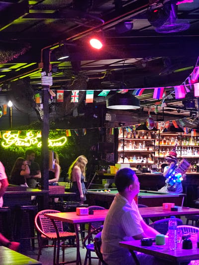The image depicts an indoor scene at a bar or restaurant, likely in Ban Klong Haeng, Thailand, characterized by low lighting and vibrant neon and colored illumination. In the foreground, a man with short dark hair wearing a light-colored, possibly white, short-sleeved collared shirt is seated at a table, looking towards his left. The table in front of him has a water bottle, a small green object, and an ashtray. To his right, a purple chair is visible.

In the midground, several individuals are present. To the left, a woman with dark hair is seen in profile, and beside her, a man in a dark shirt is also in profile. Further back, a woman with long blonde hair is facing left, wearing a dark top. To the right of the blonde woman, a pool table with balls is set up.

The background features a well-stocked bar with numerous bottles displayed on shelves illuminated by spotlights. A person, partially obscured, is standing behind the bar, appearing to interact with patrons and possibly holding a light-up device or prop. Overhanging the scene are fans mounted on the ceiling and various strings of small national flags, contributing to a festive atmosphere. Overhead, a bright red spotlight is visible, along with green neon signage that reads "Green Surprise". The overall environment suggests a lively nightlife establishment.