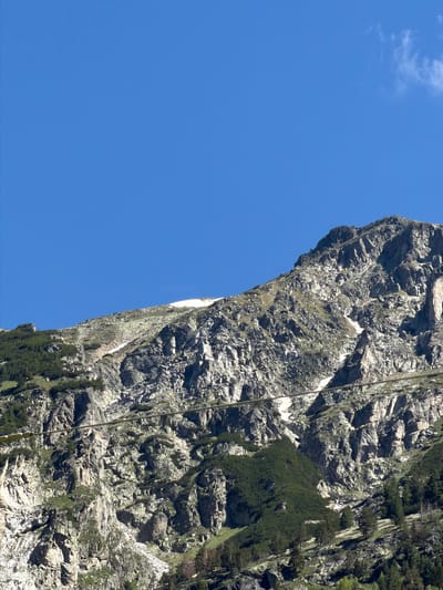 The image depicts a mountainous landscape with a clear blue sky. The lower portion of the image shows rugged, rocky slopes covered with patches of green vegetation, including trees. There are visible remnants of snow on some of the higher rocky areas. A thin, dark line, possibly a cable or wire, stretches horizontally across the middle of the frame. The overall scene is natural and unpopulated, suggesting a remote or wilderness area. The location is identified as Govedartsi, Bulgaria, a town known for its proximity to the Rila Mountains, which is consistent with the visual elements of the image.