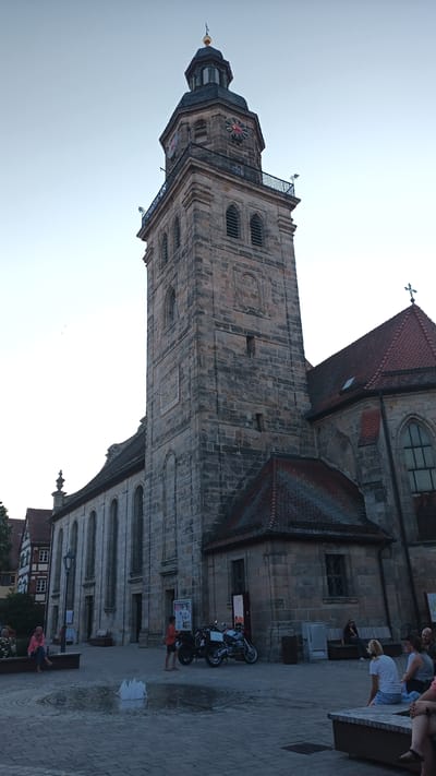 This media file depicts a bustling public square in front of a large, historic stone church. The church features a prominent bell tower with a clock and a dark, octagonal spire topped with a golden finial. The tower has multiple levels with arched windows and decorative stonework, including a sundial on one side. Adjacent to the tower is the main body of the church, characterized by long, arched windows and a red-tiled roof. A cross is visible on the roofline.

Several people are present in the square, some seated on benches and others standing. A young boy in an orange shirt stands near two motorcycles parked in the foreground. A woman in a pink hoodie sits on a bench to the left, and other individuals are visible in the background, also on benches, some appearing to be couples. A small fountain in the center of the paved square adds a dynamic element to the scene, with water jets creating a spray.

The setting appears to be Altdorf bei Nürnberg, Germany, based on the architectural style of the church and the neighboring buildings which show traditional German half-timbered construction. The lighting suggests it is either early morning or late afternoon, with the sky clear and a soft glow. The overall mood is relaxed and communal, with people enjoying the outdoor space. No discernible text is visible in the image.