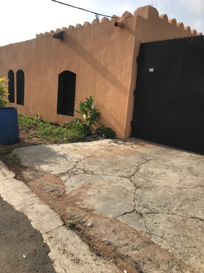 The image shows a building in Oko Erin, Nigeria. The structure is a single-story building with an arched roofline. The building is made of a light brown material and has two arched windows, one above the other. A black gate covers part of the building's facade. A small garden grows at the base of the building, and a blue barrel sits near the bottom of the frame. The concrete sidewalk and roadway are cracked and uneven. The sky is visible in the top left corner of the image.