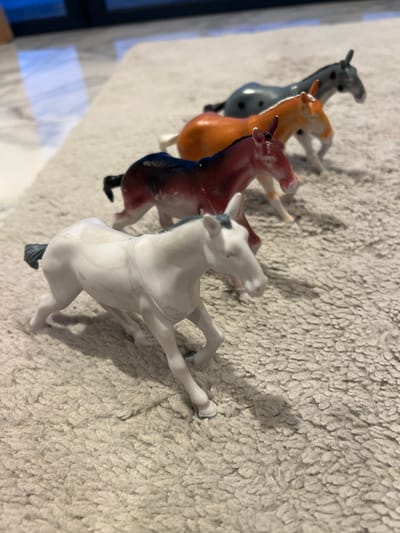 Four toy horses are lined up on a textured, light-colored rug. The horses are positioned from closest to furthest from the camera, and they appear to be in motion. The closest horse is white with dark grey markings on its mane and tail. The next horse is a reddish-brown with black markings. The third horse is orange with black spots. The furthest horse is grey with black spots. The background shows a blurred reflection of a blue and white tiled floor and a dark vertical object, possibly a doorway or furniture. The scene is indoors, with soft lighting. There is no indication of Baan Harn, Thailand.