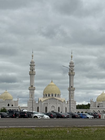 The image captures the White Mosque in the city of Bolgar, Republic of Tatarstan, Russia, under a heavily overcast sky. The grand white structure features multiple domes, the largest of which is golden, and two prominent minarets flanking the central building. The mosque's architecture is intricate, with detailed carvings and arched windows. In the foreground, a line of cars is parked. These vehicles include a black SUV with a roof rack, several sedans in black, white, and blue, and a red car visible at the far right. A paved road separates the parking area from a strip of green grass. The background shows a dense line of trees behind the mosque. The overall atmosphere is somber due to the cloudy weather.
