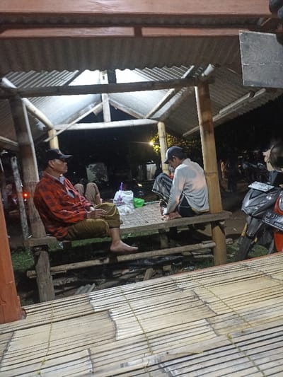 The image depicts an evening scene under an open-air structure in Kecamatan Pujut, Indonesia. The time is clearly night, illuminated by artificial lights casting shadows.
In the foreground, two men are prominently seated on a raised wooden bench, both barefoot. The man on the left wears a red and black plaid shirt, brown pants, and a dark baseball cap, looking towards the left side of the frame. The man on the right wears a light-colored long-sleeve shirt, striped lower garment (possibly a sarong or pants), and a dark baseball cap, looking down. Another person is partially visible behind the man in plaid.
The structure itself is constructed with bamboo poles and features a corrugated metal roof. Below the seating area, the ground appears to be dirt with some scattered debris. A bamboo-slatted platform or floor extends into the foreground, showing signs of wear.
Between the two main figures, on the bamboo platform, a white plastic bag displaying the text "TARA" and a green plastic bag are visible. To the far right, a dark-colored scooter motorcycle is parked, with part of its license plate visible, reading "DP 3710 Q" and "08 24". In the background, more people are dimly visible, suggesting a small gathering or communal space. The overall atmosphere appears relaxed and informal.