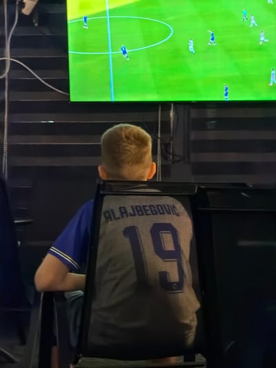 The image shows a young person from behind, wearing a blue and white striped t-shirt and a grey jersey with the name "ALAJBBEGOVIĆ" and the number "19" printed on the back. The person is seated and facing a large television screen that displays a football match. The screen shows players on a green field. The setting appears to be indoors, with dark, striped blinds visible behind the person and to the right of the television. The person's blonde hair is visible above the jersey. The overall lighting suggests an indoor environment with the television being the primary light source. The location is specified as Nević Polje, Bosnia and Herzegovina.