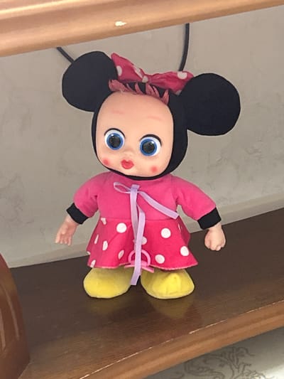A doll with large black ears and a red and white polka-dotted bow is standing on a wooden shelf. The doll is wearing a pink dress with a white polka-dotted skirt and a purple ribbon tied around its waist. Its face is round with large blue eyes and red lips. The doll's feet are yellow and rounded. The background appears to be a beige wall with a subtle texture. The image is a medium shot of the doll, focused on its upper body and legs. The scene is indoors and appears to be a domestic setting. The doll is the sole subject in the image. There is no information available about the location in Buynaksk, Russia.