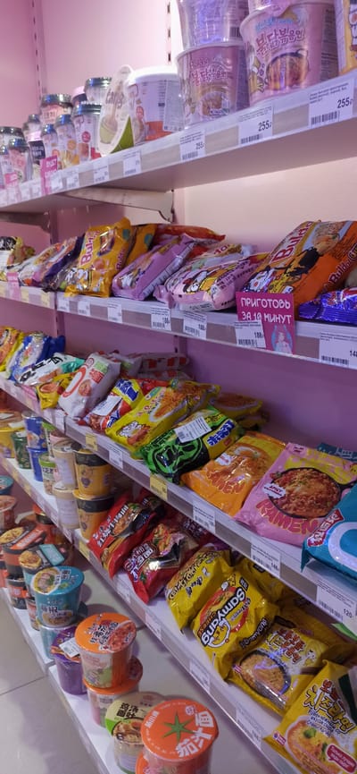 The image shows a shelf in a store, likely a supermarket, displaying various types of instant noodles and snacks. The products are arranged on white shelves with pink backlighting. Several brands of instant noodles are visible, including Samyang, Buldak, and others with packaging in Korean and Chinese. Some of the products are in cups, while others are in pouches. Prices are displayed on some of the items, ranging from 188 rubles to 255 rubles. The store appears to be well-stocked, with many items filling the shelves. The overall impression is a retail environment specializing in Asian food products. The provided information states that this location is in Asbest, Russia. There are no people visible in the image.