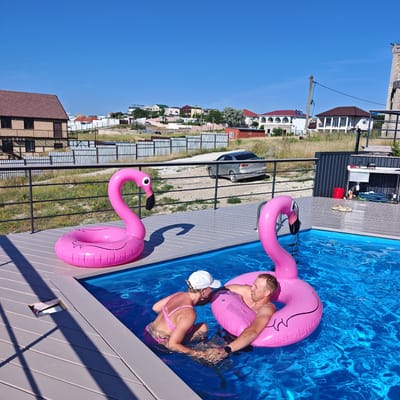 Two people are relaxing in a bright blue swimming pool on a sunny day. They are both using large, inflatable pink flamingo pool floats. The woman is wearing a pink bikini and a white baseball cap, and the man is wearing a watch on his left wrist. They are leaning against each other, appearing to be in a comfortable and friendly interaction.
The pool is surrounded by a grey wooden deck, and a black metal railing runs along the edge. In the background, there is a dry, grassy area with several houses, fences, and a silver car. The sky is a clear, deep blue, suggesting it is a warm and pleasant day. The overall setting appears to be a residential area, possibly a backyard or vacation property. There is some visible text on the inflatable flamingo floats, but it is too blurry to read. The scene evokes a sense of leisure and enjoyment.