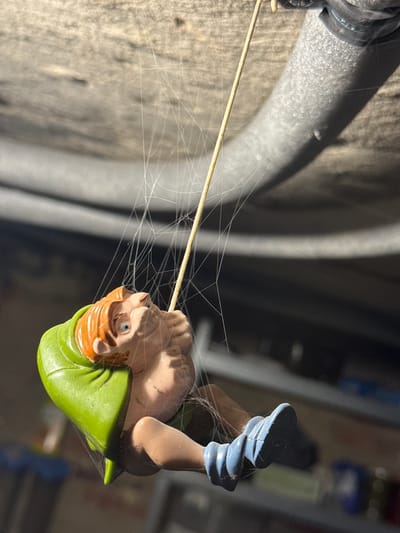 A close-up, low-angle shot captures a small plastic figurine of a character with orange hair, blue eyes, and a green hooded cape, resembling Quasimodo from Disney's "The Hunchback of Notre Dame." The figurine is suspended mid-air, entangled in a prominent spiderweb that stretches across the frame. The character is depicted in a dynamic pose, with arms outstretched and legs bent, appearing to be falling or swinging. The figurine is wearing blue shoes. The background is out of focus, revealing the textured surface of what appears to be a wooden ceiling or beam above, and a dark, indistinct area below, suggesting an indoor or sheltered environment like a garage or attic. A grey, insulated pipe runs diagonally across the upper right portion of the image. The lighting creates strong shadows and highlights, emphasizing the delicate strands of the spiderweb and the textured surfaces. The location is not discernible from the image alone.