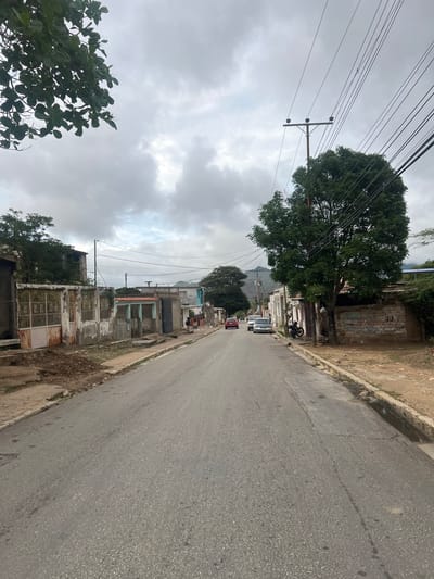 The image depicts a street scene in El Maco, Venezuela, under an overcast sky. The paved road, showing some signs of wear and tear, stretches towards the background. On the left side of the road, there are various buildings with weathered facades and protective metal gates. Some areas show exposed earth and debris. On the right side of the road, there are more buildings, some with a more rustic appearance, and a large tree with dense foliage. Several cars are parked or driving on the street, including a white sedan and a red car further down the road. A motorcycle is parked near the large tree. Power lines are visible overhead, supported by a utility pole. The background shows more buildings and some hilly terrain with vegetation. A person is visible walking on the sidewalk on the right side of the street.