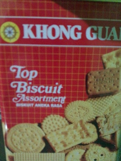 The image shows a close-up of a package of “Khong Guan” biscuits, with the text “Top Biscuit Assortment” in white letters on a red background with a grid pattern. The package is located at the coordinates -5.45 latitude and 105.313 longitude, which is in the city of Bandar Lampung, Indonesia. The image is taken at a slight angle, revealing multiple assorted biscuits on the bottom of the package. The biscuits are arranged in a layered formation, with a variety of shapes and sizes. The image is focused on the packaging and biscuits and does not show any people or other objects. The environment is not visible.
