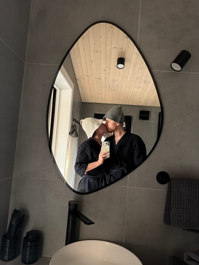 A selfie of two people kissing is reflected in an irregular-shaped mirror in a bathroom. The bathroom is predominantly gray with light wood accents. One person wears a gray beanie and a dark waffle-textured robe, holding a phone to take the picture. A sink, hair dryer, and towels are visible in the bathroom. The location is Капцёўскі сельскі Савет, Belarus.