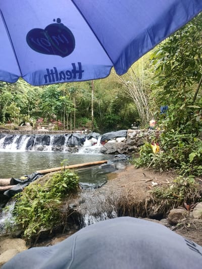An outdoor scene depicts a small, man-made waterfall cascading into a pool of water. The area appears to be a natural setting with lush green vegetation and bamboo trees in the background. A blue umbrella, with a logo featuring two heart shapes and the word "Health" written in reverse, is positioned in the foreground, partially obscuring the view. Several people are visible near the waterfall, some appearing to be bathing or relaxing. Objects such as black tarps, rocks, and bamboo logs are present, suggesting a rudimentary setup for recreational purposes. The ground is muddy and covered with vegetation and debris. The overall impression is a simple, informal recreational spot. There is no visible indication of the specific location within Kidapawan City, Philippines, beyond it being an outdoor, natural setting.