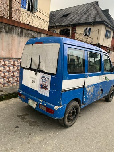 A blue and white minibus is parked on the side of a street. The vehicle has visible wear and tear, with scratches and chipped paint. A poster, featuring people and the words "JERICHO MARCH," is attached to the back of the van. The back window is covered with a white sheet. A building with a dark grey roof is visible in the background, along with a fence topped with barbed wire. The road is gray and shows some signs of wear.