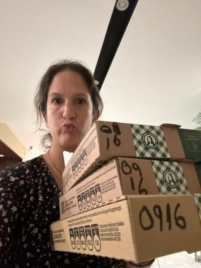 In San Isidro, Peru, a woman with dark hair and pursed lips holds a stack of four small cardboard boxes. The boxes are stacked diagonally, with the topmost box angled more sharply than the others. The woman wears a black dress with a floral pattern and a thin gold necklace. Black handwritten numbers are visible on the sides of the boxes: "09 16" is written on the top box and on another box below it. The boxes have green and white checkered logos with a woman's profile on them. Text on the boxes appears to be in Spanish and includes nutritional information. The background is out of focus, but shows a wall and a ceiling with a light fixture.