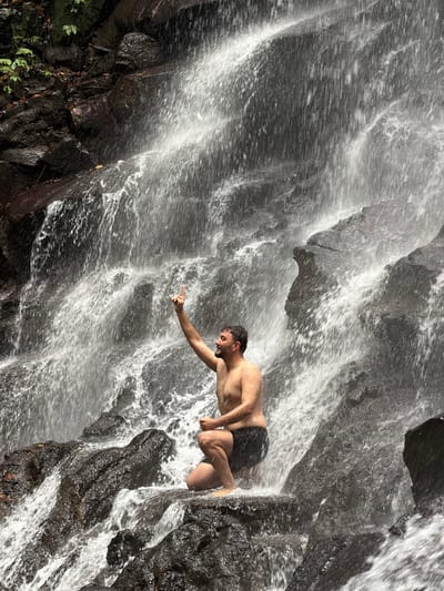 A shirtless man is kneeling on dark, wet rocks in front of a waterfall. He is wearing black swim trunks and has his left arm raised with his index finger pointing upwards. The waterfall cascades down a rocky cliff face, with white water creating a misty effect. The surrounding environment is lush and green, suggesting a natural, tropical setting. The image appears to be taken in the city of Gianyar, Indonesia, a region known for its numerous waterfalls.