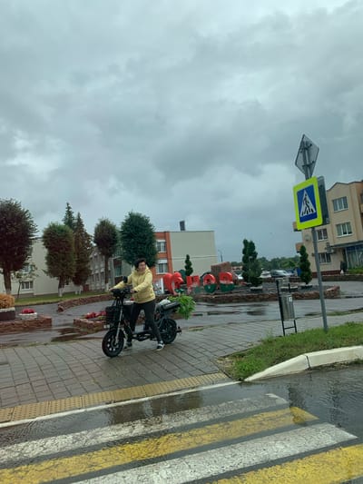 In Snovski Sel'skiy Sovet, Belarus, on an overcast day, a woman rides a black electric scooter equipped with a basket. She is wearing a yellow hooded jacket and dark leggings. The scooter is on a wet, paved sidewalk, indicated by the reflective surface and puddles. To her right, there is a pedestrian crossing with yellow and white stripes, also partially submerged in water. Further into the background, a large, colorful art installation with Cyrillic letters is visible. Behind the installation, there are trees and buildings, including a multi-story building with orange and beige facades. A pedestrian crossing sign is mounted on a pole to the right of the installation. The overall environment suggests a town square or a public area that has experienced recent rainfall.
