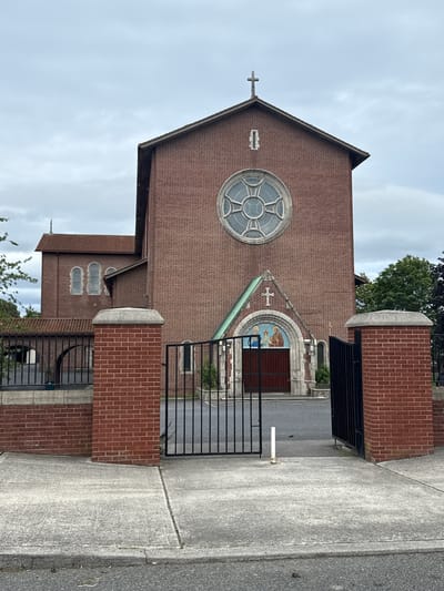 The image shows the exterior of a brick church. The church has a tall, peaked roof with a cross at the apex of the facade. A large, circular stained-glass window with a geometric pattern is visible on the upper part of the facade. Below this, an arched entryway is decorated with a mosaic depicting religious figures. A narrower section of the church to the left has arched windows. The church is bordered by a sidewalk and a black metal fence supported by brick pillars. The sky is overcast. The image is oriented vertically. No people or events are depicted. The context provided is Dublin, Ireland.
