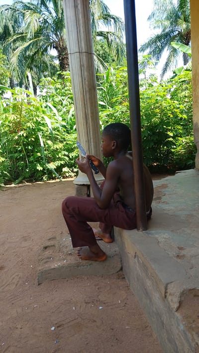 A young boy, shirtless and wearing maroon pants, sits on a concrete step outdoors. He is focused on a knife he holds in his hands, appearing to be whittling or working on an object. The boy's bare feet rest on the step and the sandy ground in front of him. He is in profile, facing towards the right of the frame.
Behind the boy, lush green vegetation, including palm trees and leafy plants, dominates the background. A large, fluted concrete column stands to his left, and a dark metal pole is positioned to his right, partially obscuring his body. The ground is covered in reddish-brown sand with scattered small white debris. The scene appears to be in a tropical or subtropical climate, with bright daylight suggesting it is daytime. The overall mood is one of quiet concentration. No text is visible in the image. The location is Donga, Nigeria.