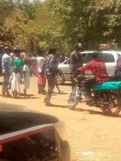 The image depicts an outdoor scene in Kagoma, Nigeria, under bright daylight on an unpaved, light brown surface. Approximately twenty individuals are gathered, comprising adults and children. In the left-center, a person in a dark camouflage uniform with a dark cap stands facing left, positioned near a group of civilians. This group includes a child in a white dress and another child in a light patterned outfit. A small white child's bicycle lies on its side on the ground near these civilians. To the right, a male individual wearing a red and black plaid shirt, black trousers, and pink sandals is seated on a black motorcycle with chrome accents, facing right, and wearing a black helmet. Behind the motorcycle, a white SUV, possibly an Opel Zafira, is parked with luggage secured on its roof rack. A man in a dark shirt and a black baseball cap stands near the front of the white SUV, facing the motorcycle rider. Dense green foliage and trees form the background. A partial view of a black vehicle is visible in the immediate foreground, showing its front passenger side headlight and a red and white stripe along the hood.
