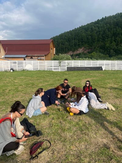 A group of people are sitting and lying on the grass in a rural outdoor setting. They appear to be at a gathering or event, possibly a festival, given the relaxed atmosphere and casual attire. The scene is set against a backdrop of a wooded hillside with a clear sky overhead, suggesting a daytime event. A white wooden fence runs along the edge of the grassy area, and a building with a reddish-brown roof is visible in the distance. Some individuals are looking at their phones, while others are engaged in conversation or simply resting. The overall mood seems peaceful and informal. The time of day appears to be afternoon, with the sun casting shadows on the grass. The location is identified as Solontsy, Russia. There is no visible text in the image.